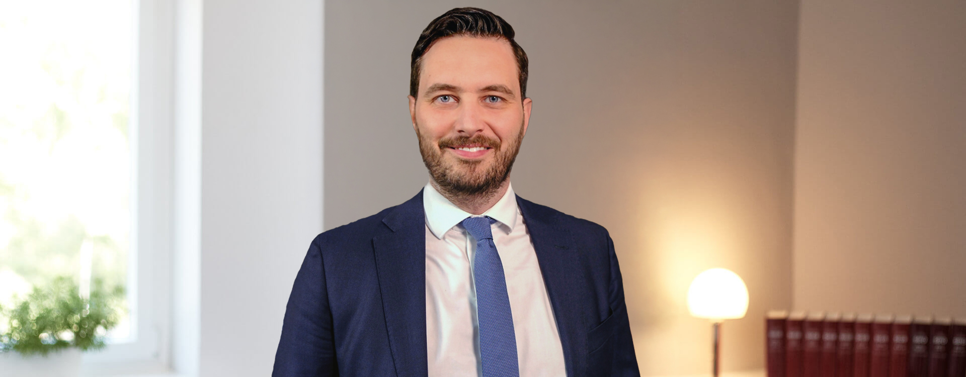 Dr. Frédéric Schneider, SCHNEIDER || MICK Strafverteidigung, im dunkelblauen Blazer im hellen Kanzleibüro mit Bücherregal
